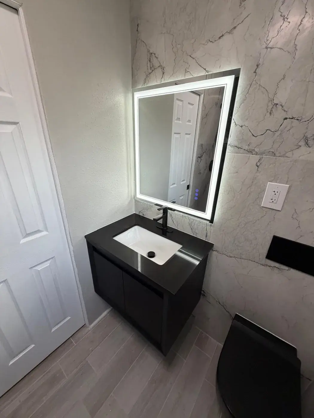 Powder room with a floating black vanity, a backlit mirror and marble-look wall tile.