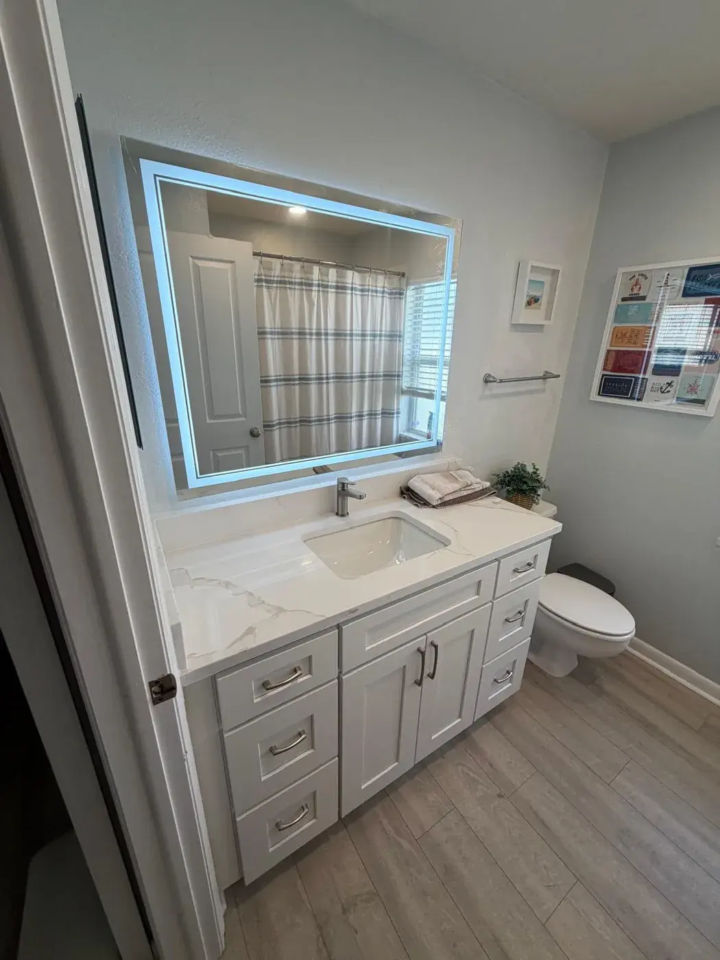 Bathroom vanity with white shaker cabinets, a quartz top, an undermount sink, a lit wall mirror and wood-look floor tile.